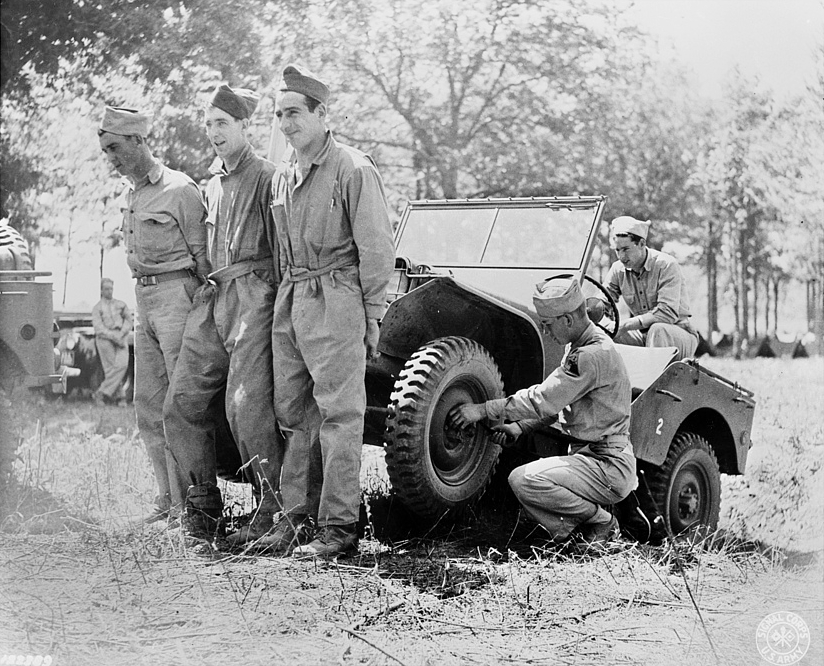 World War Two-era black and white photogrpah of three soldiers holding up the front of a Jeep, while a fourth soldier changes the front tire.